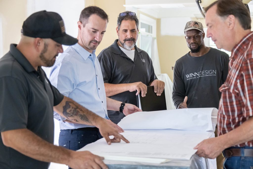 Five men stand around a table looking at blueprints. Two men in dark shirts point at the plans, while the others, dressed casually, watch attentively. The setting appears to be a construction or renovation site with light walls and covered furniture.