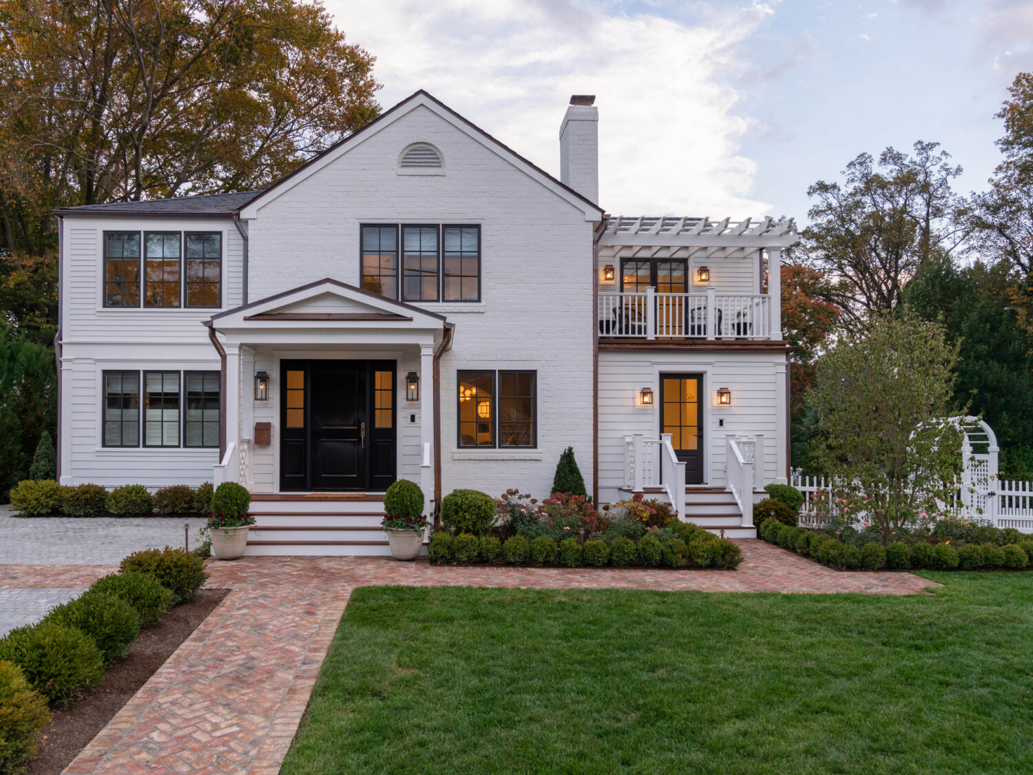 A two-story white brick house with black window frames, double front doors, two staircases leading to entrances, a balcony with a pergola, manicured bushes, a brick walkway, green lawn, and trees in the background at sunset.