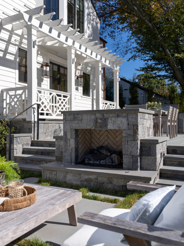 A sunlit outdoor patio features a stone fireplace with herringbone brickwork, steps leading up to a white house with a pergola, and a dining table. Cushioned seating and a wooden table with decor are in the foreground. Trees and a clear blue sky are visible.