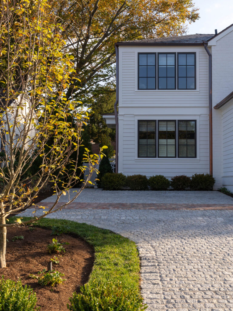 A modern white house with black-framed windows sits behind a cobblestone driveway. In the foreground, there is a curved pathway bordered by green grass and a landscaped garden bed with a young tree. The scene is lit by soft, late-afternoon sunlight.