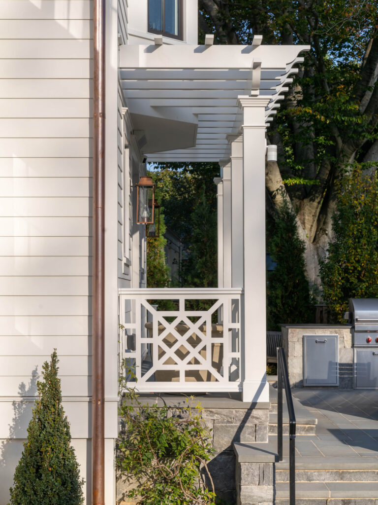 A side view of a white house porch with a decorative pergola, crisscross wooden railing, stone steps, and a lantern. Shrubs and a tree are visible, along with an outdoor grill and built-in countertop in the background.