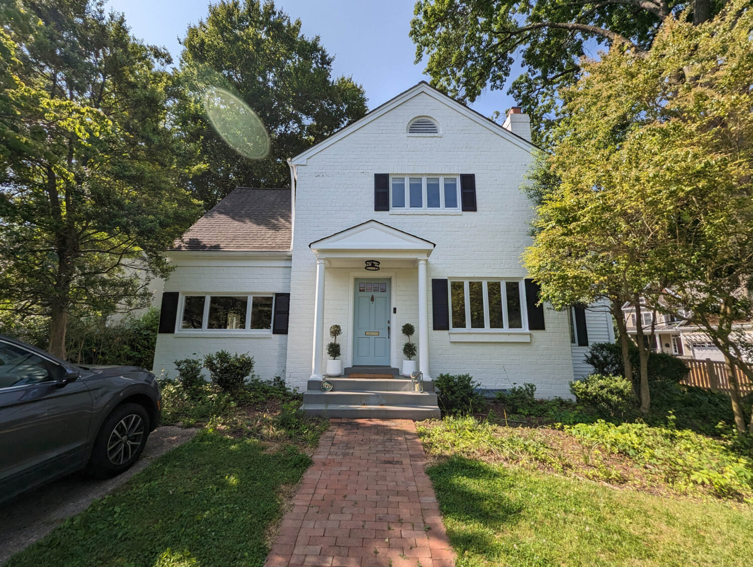 A white, two-story house with black shutters and a small front porch stands among green trees. A brick path leads to the blue front door with potted plants on each side. A gray car is parked to the left, and sunlight shines through the trees above.